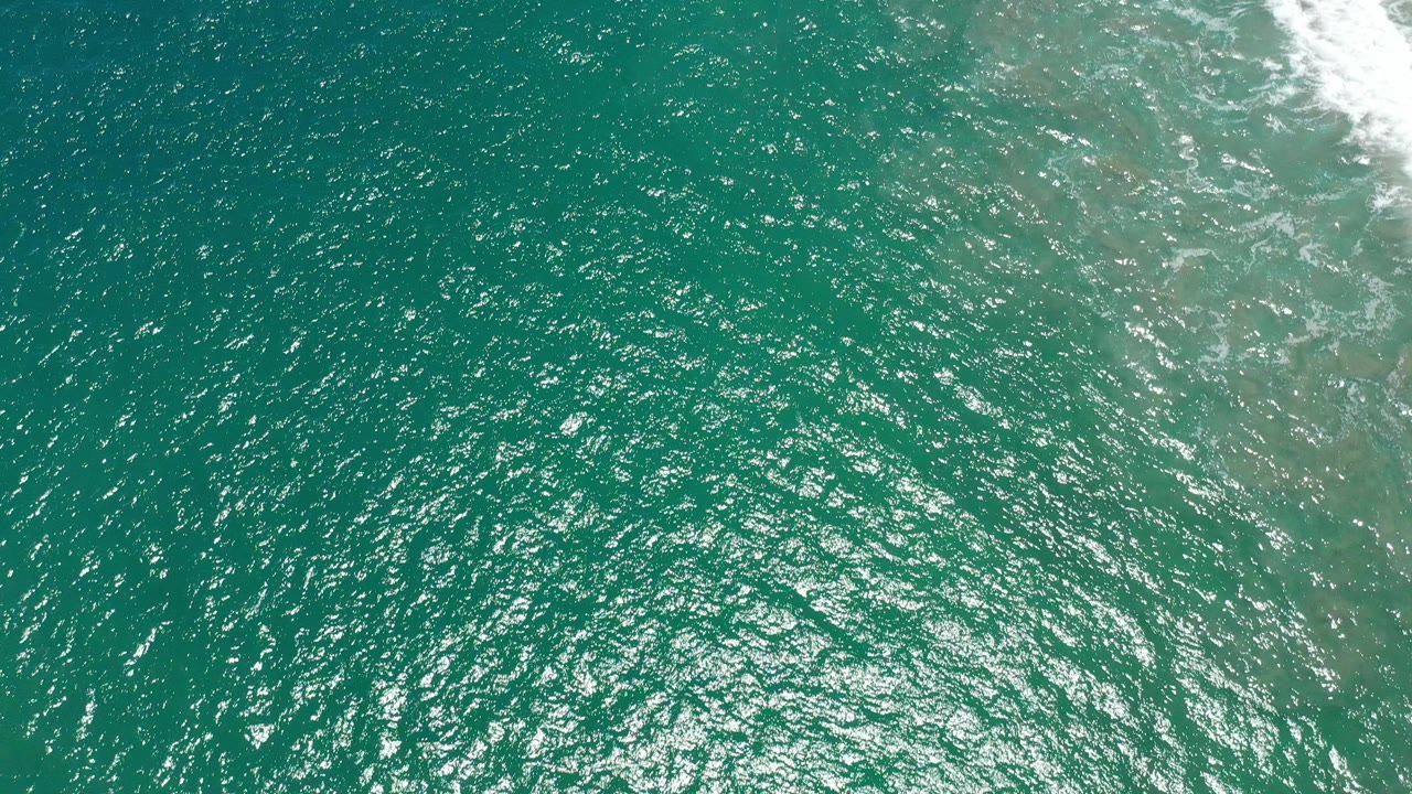 Top-down view of green-blue ocean water in San Jose del Cabo, Mexico, with sunlit ripples, pale underwater color near the right edge, white surf at the top right, and no visible people.