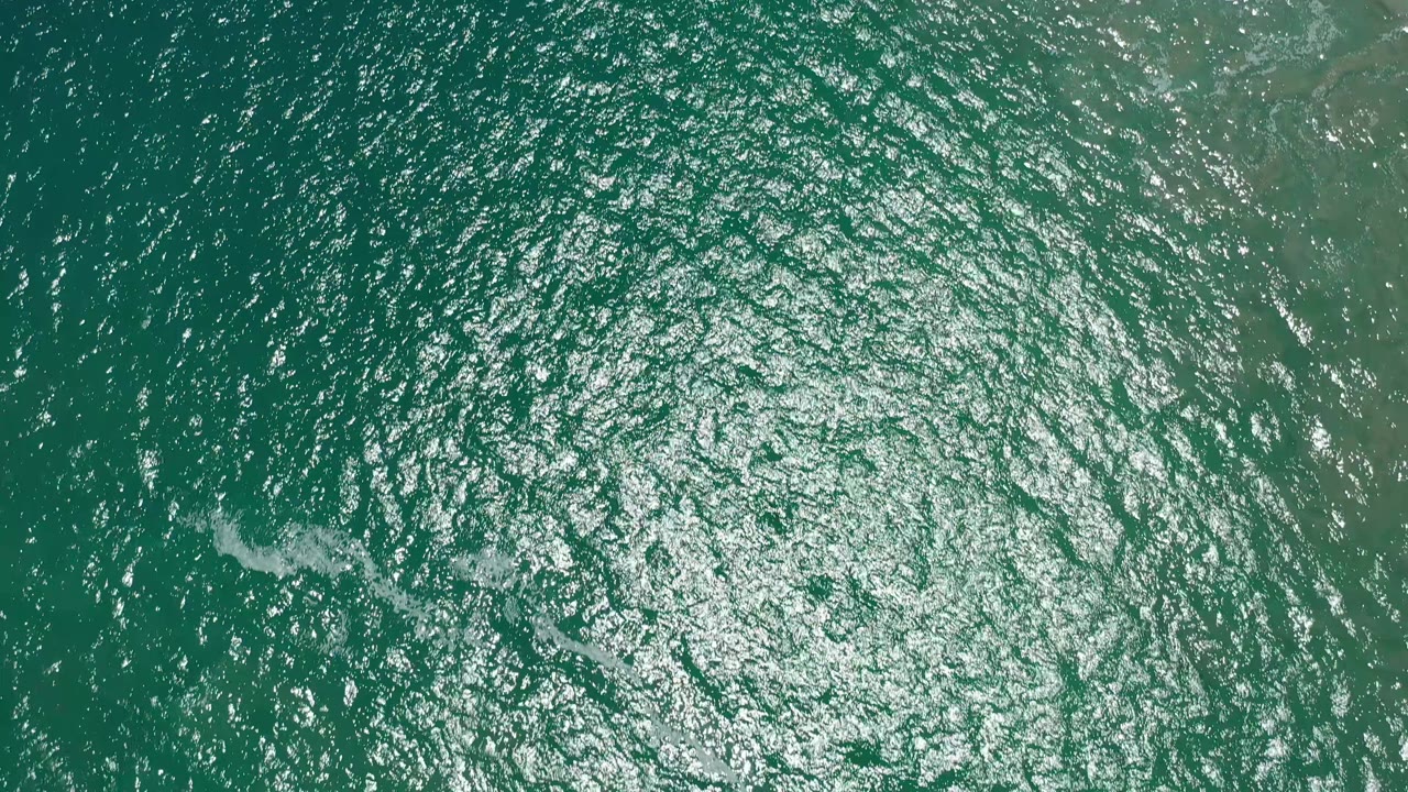 Top-down San Jose Del Cabo water frame with sun glitter spread through the center and a small pale wave streak left of center, no visible people.