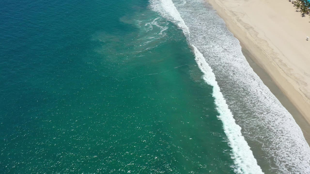 Aerial San Jose del Cabo coastline frame with more open turquoise water filling the left side, foamy white surf crossing diagonally, pale beach to the right, and no clearly visible people.