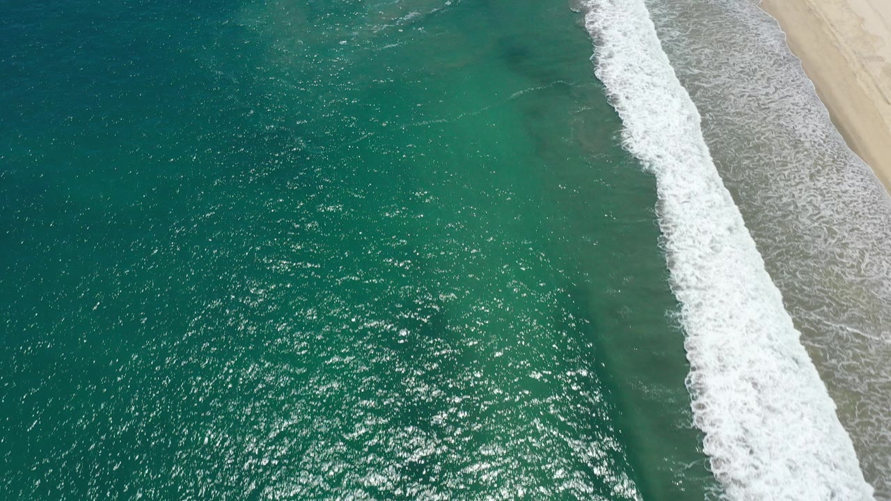 Overhead view of turquoise water and a diagonal white wave line meeting pale sand in San Jose del Cabo, Mexico, with no visible people.