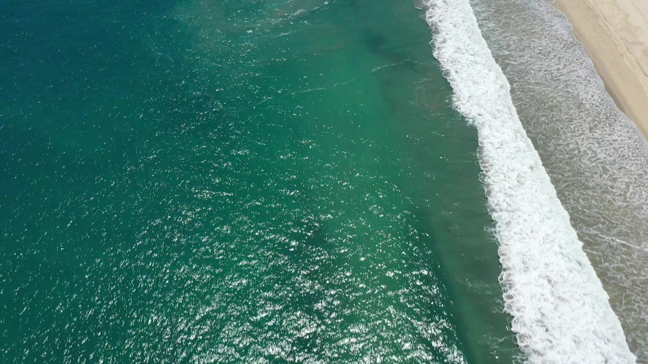 Top-down San Jose del Cabo shoreline frame with sunlit green-blue water, a thicker band of white foam, pale beach at the right edge, and no visible people.