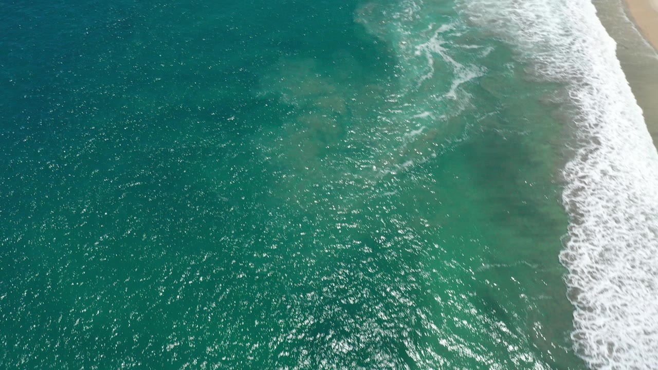 Overhead view of turquoise water and white surf meeting a sandy shoreline in San Jose del Cabo, Mexico, with sunlight on the water and no visible people.