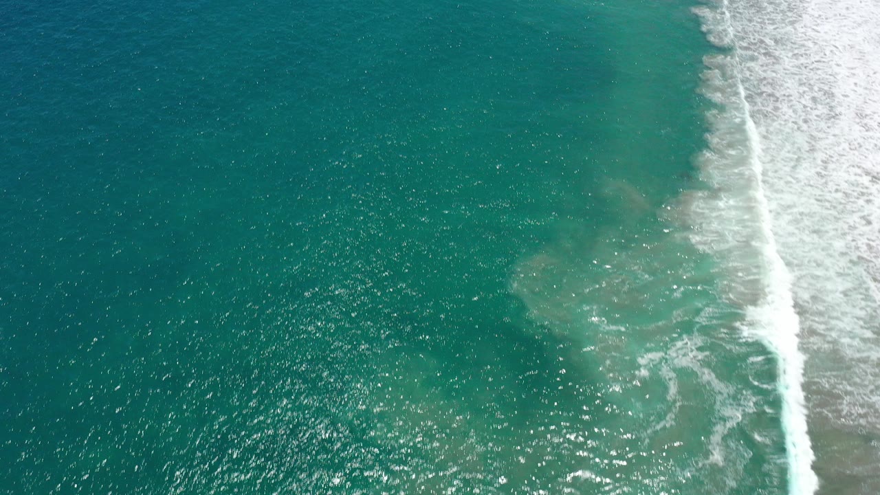 Top-down aerial view of blue-green ocean in San Jose del Cabo, Mexico, with a white wave breaking along the right edge and no visible people.