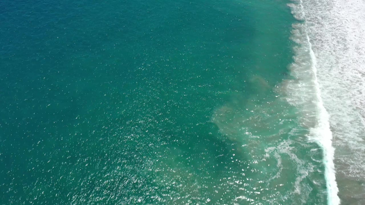 Aerial frame over San Jose del Cabo ocean water, with sunlight glittering on blue-green ripples and a bright band of surf foam at the right, no visible people.