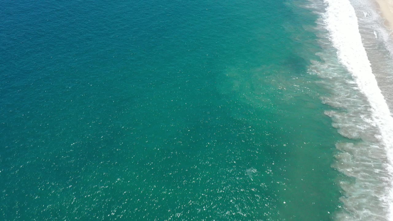 Aerial view of turquoise ocean beside a sandy shoreline with white surf.
