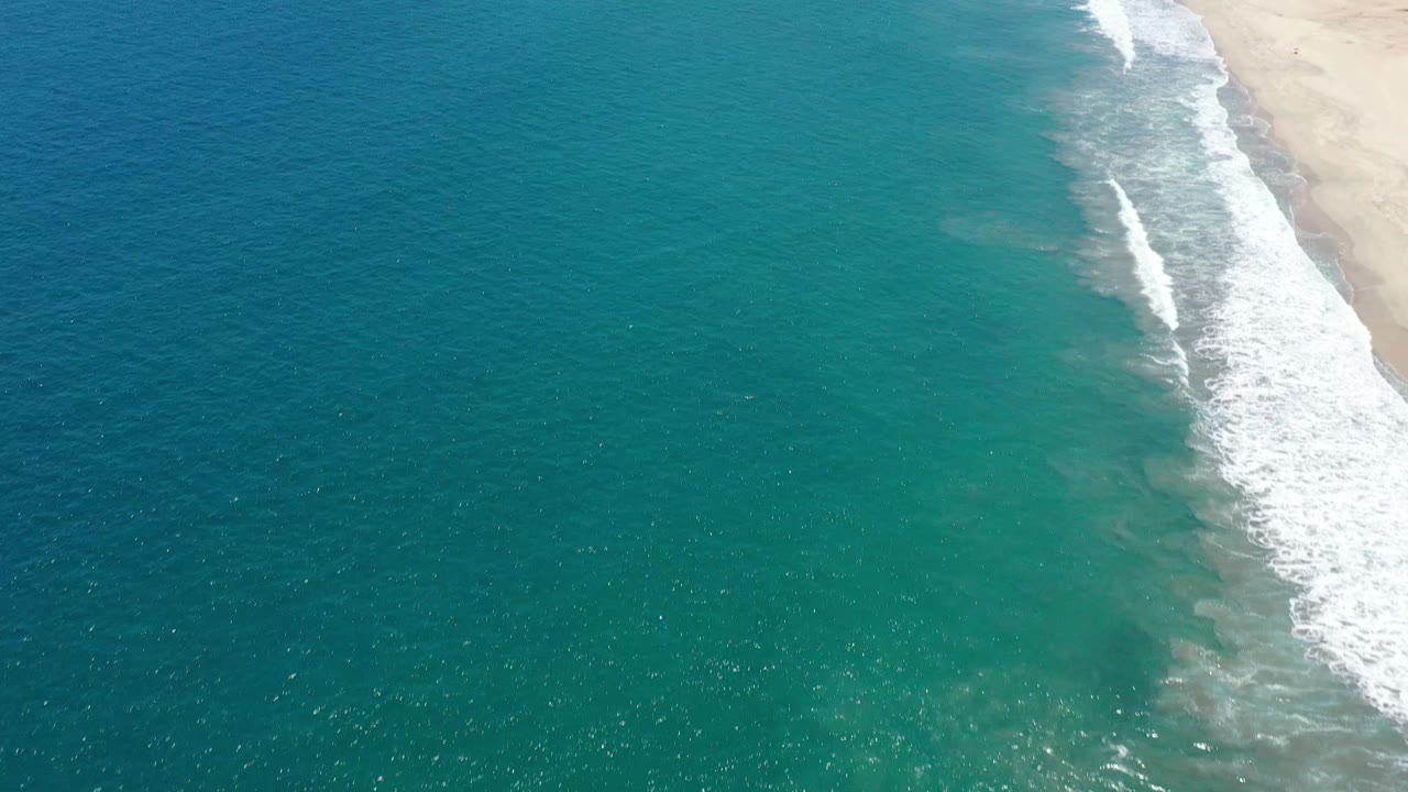 Aerial coastal frame in San Jose Del Cabo, Mexico, showing more open blue-green water, foam lines near the right edge, pale beach sand, and no visible people.