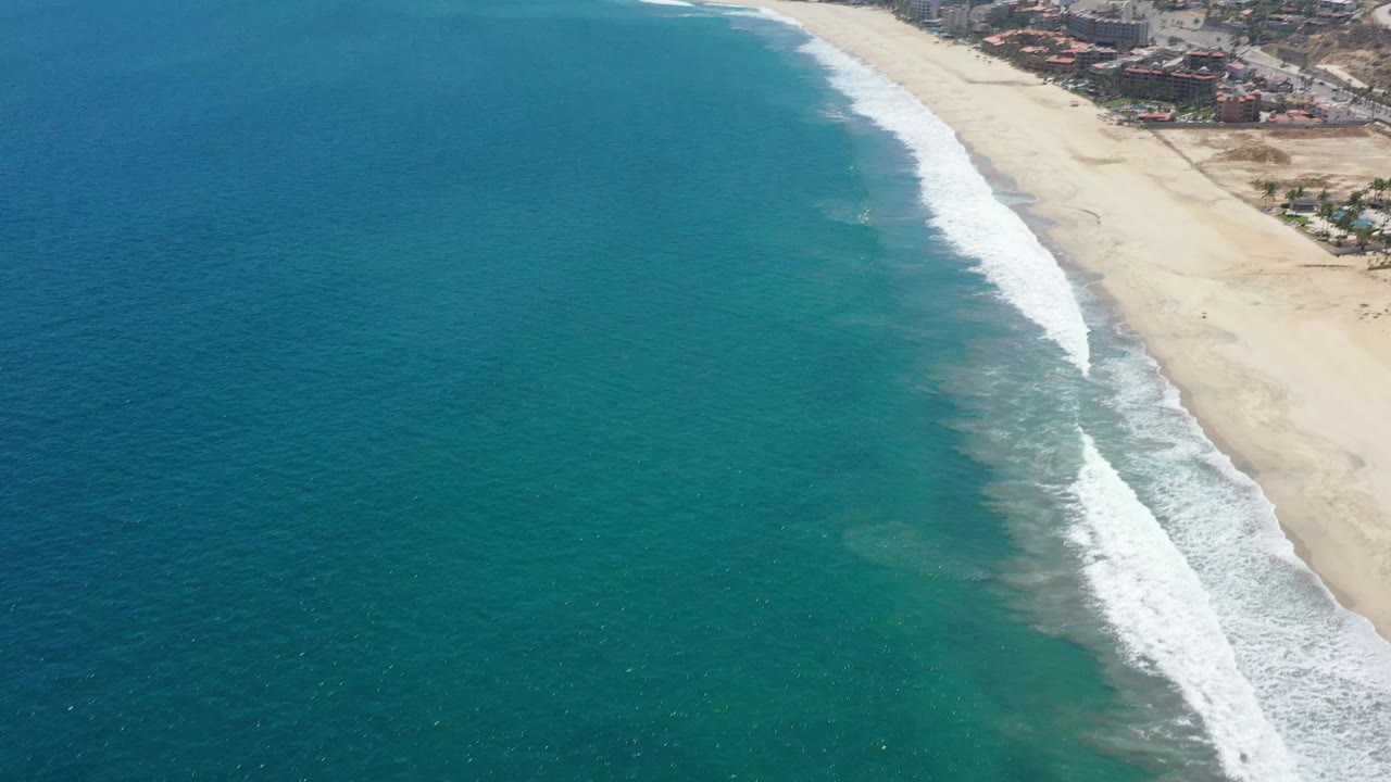 Aerial frame over San Jose del Cabo showing broad blue water, a long pale beach, a curling band of white waves, shoreline buildings, and no visible people.