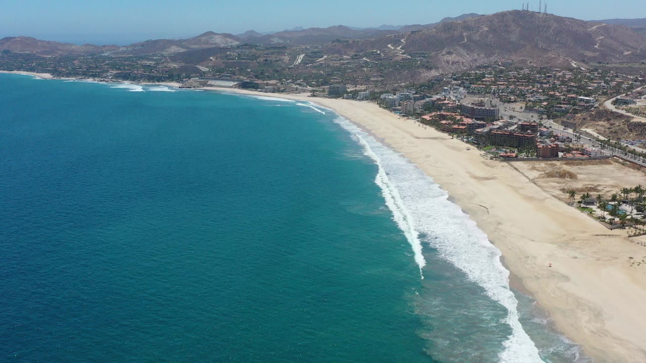 High aerial view over San Jose del Cabo, Mexico, with turquoise water on the left, a broad sandy beach, white surf, inland buildings, and no visible people.