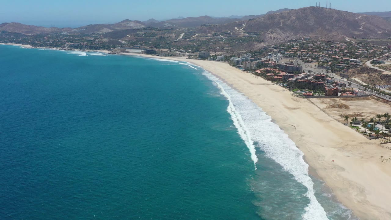 Aerial frame in San Jose del Cabo, Mexico, looking along the beach as a long white surf band curves beside blue-green water, with no visible people.
