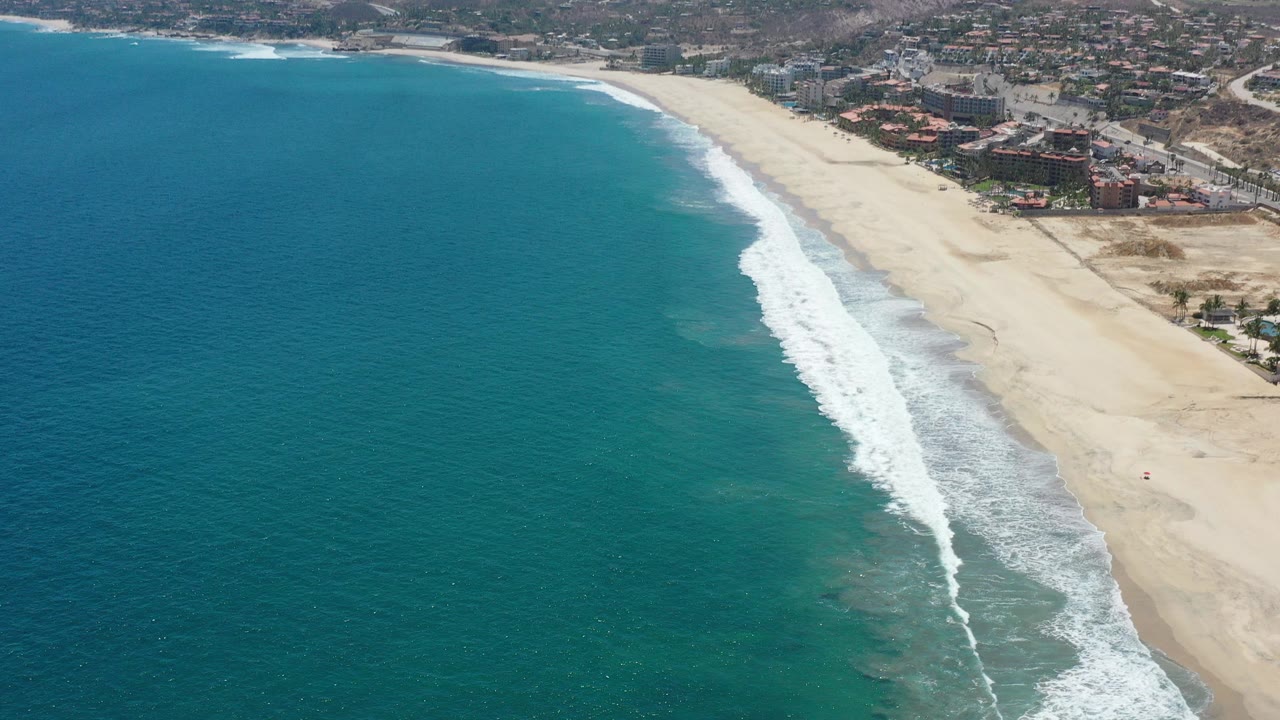 Aerial top view over San Jose del Cabo shoreline with teal-green ocean on the left, white surf crossing the sand diagonally, resort buildings in the distance, and no visible people.