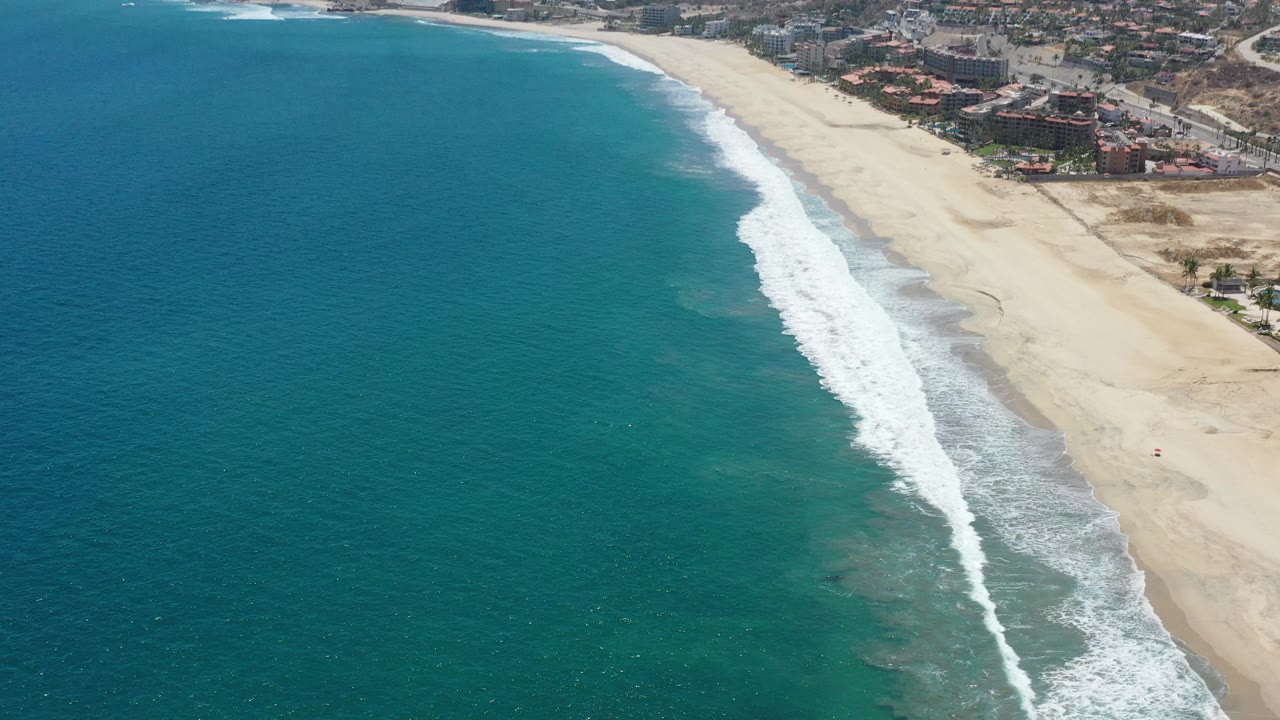Aerial coastline view with rough white waves and beachfront development beyond the sand.