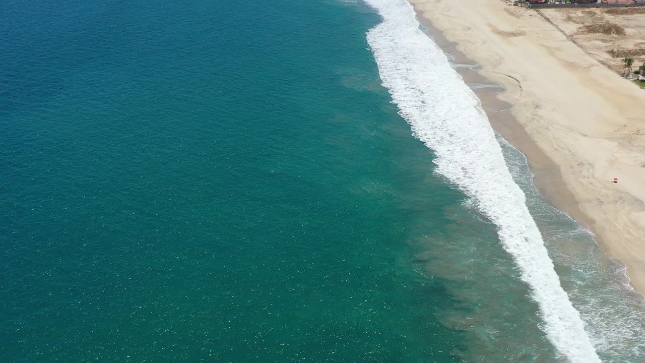 Aerial frame over the San Jose del Cabo shoreline, with teal ocean on the left, a diagonal band of white surf, pale sand on the right, and no visible people.