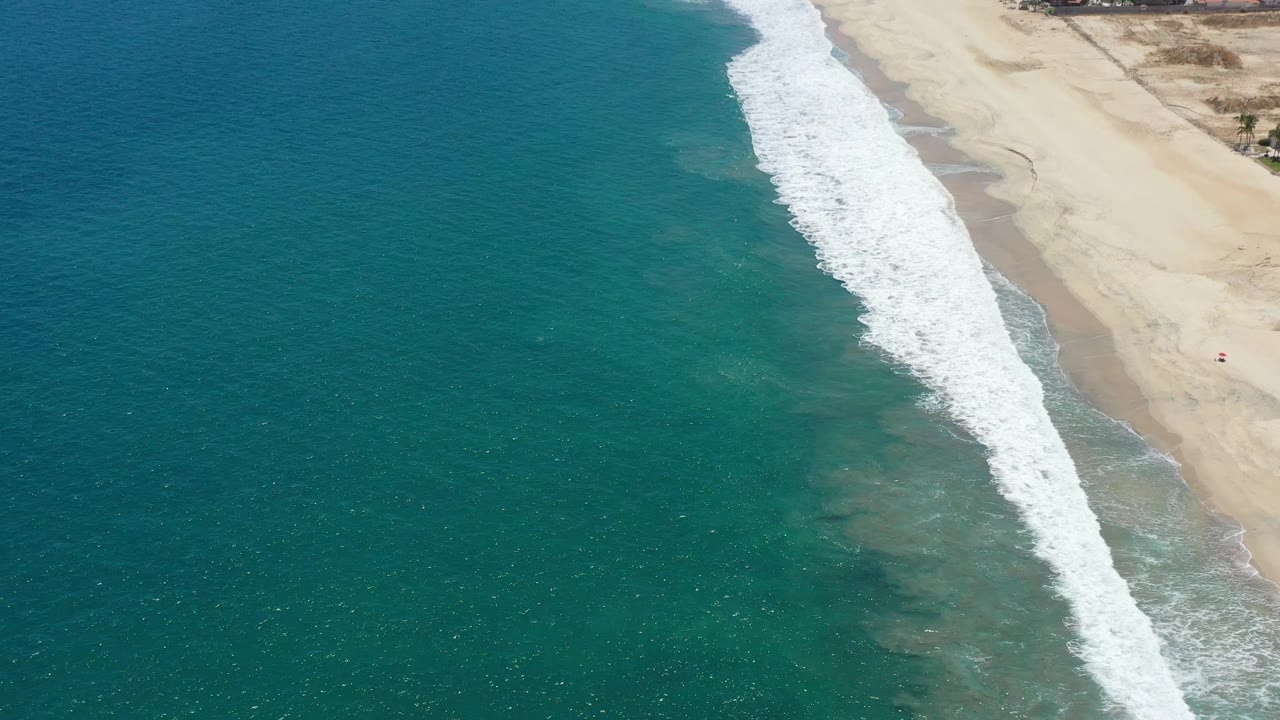 Aerial view of San Jose del Cabo surf, with the foam line shifted along the sand and broad teal water filling most of the frame, no visible people.