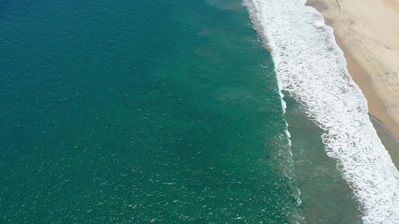 Aerial frame over San Jose del Cabo showing teal water, a diagonal strip of white surf, pale sand at the right edge, and no visible people.