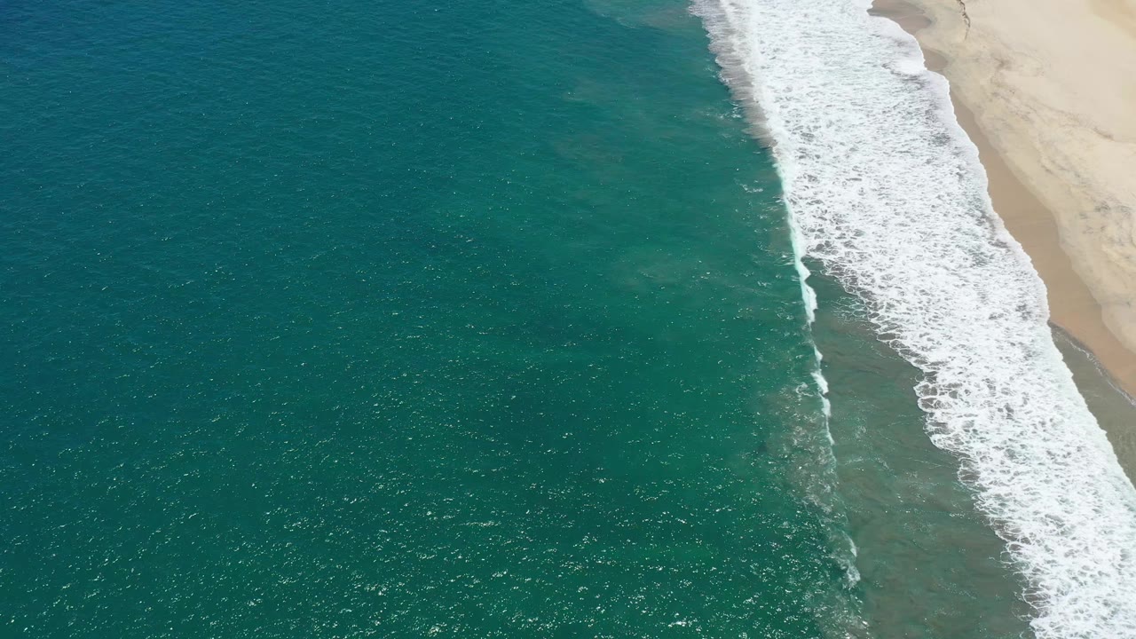 Top-down aerial frame in San Jose del Cabo with white foam spreading along the right-side beach and deeper teal water across the left, with no visible people.