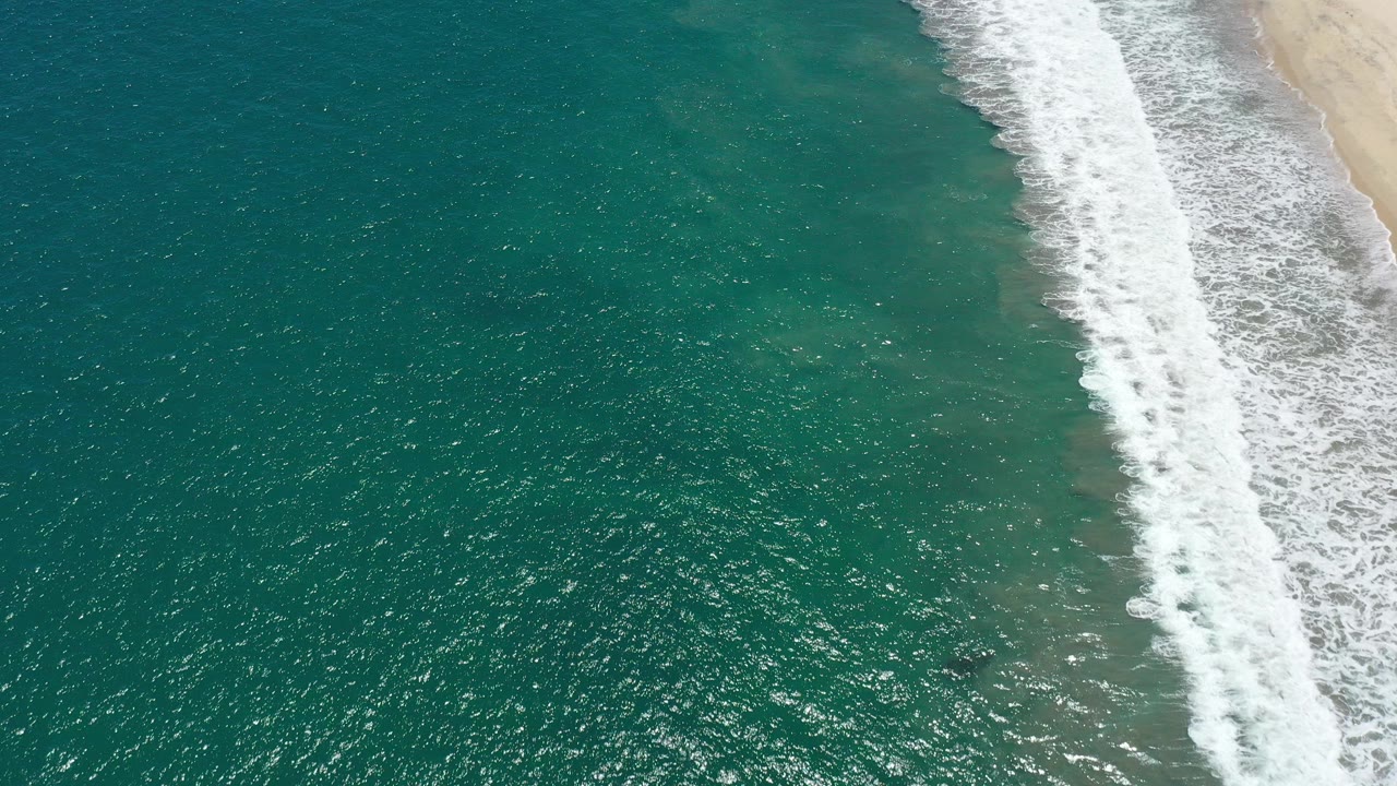 Overhead view of teal water and a diagonal strip of white surf along the sandy shore in San Jose Del Cabo, Mexico, with no visible people.