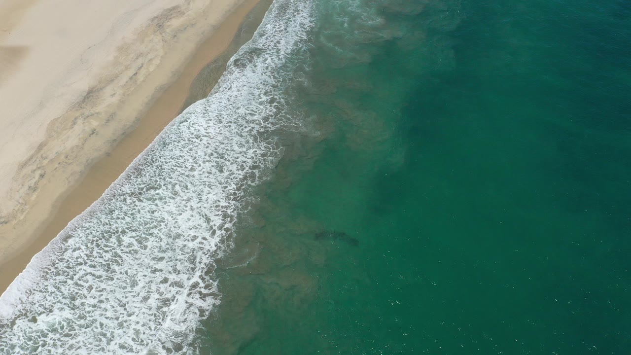 Aerial view in San Jose Del Cabo, Mexico, with tan beach at left, white surf crossing diagonally, green ocean to the right, and no visible people.