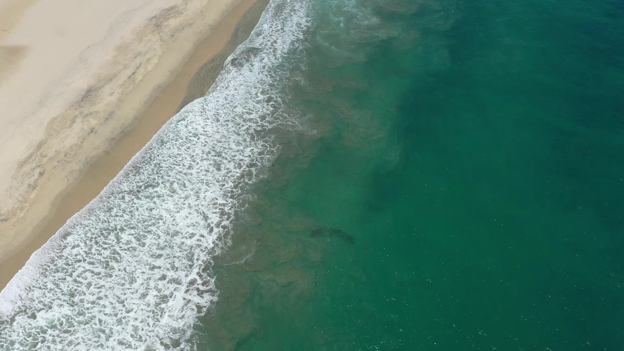 A slightly later aerial frame shows the foamy surf band shifted along the sand in San Jose Del Cabo, Mexico, with open green water and no visible people.
