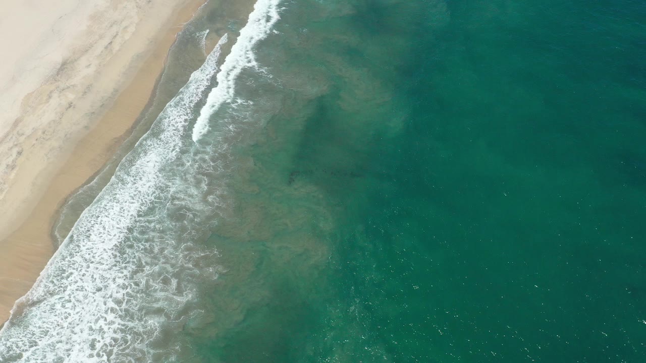 Overhead view of the San Jose del Cabo shoreline at the start of the clip, with pale sand on the left, white surf crossing diagonally, teal ocean water on the right, and no visible people.