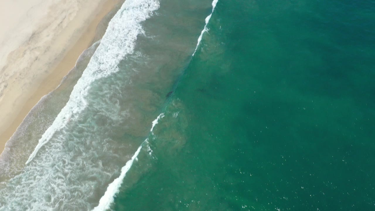 Overhead view of San Jose del Cabo shoreline with pale sand at left, teal ocean at right, diagonal white surf, and no visible people.