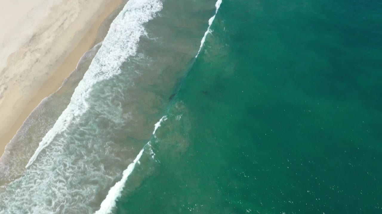 Aerial frame from San Jose del Cabo showing white foam widening along the wet sand while darker teal water fills the right side, with no visible people.