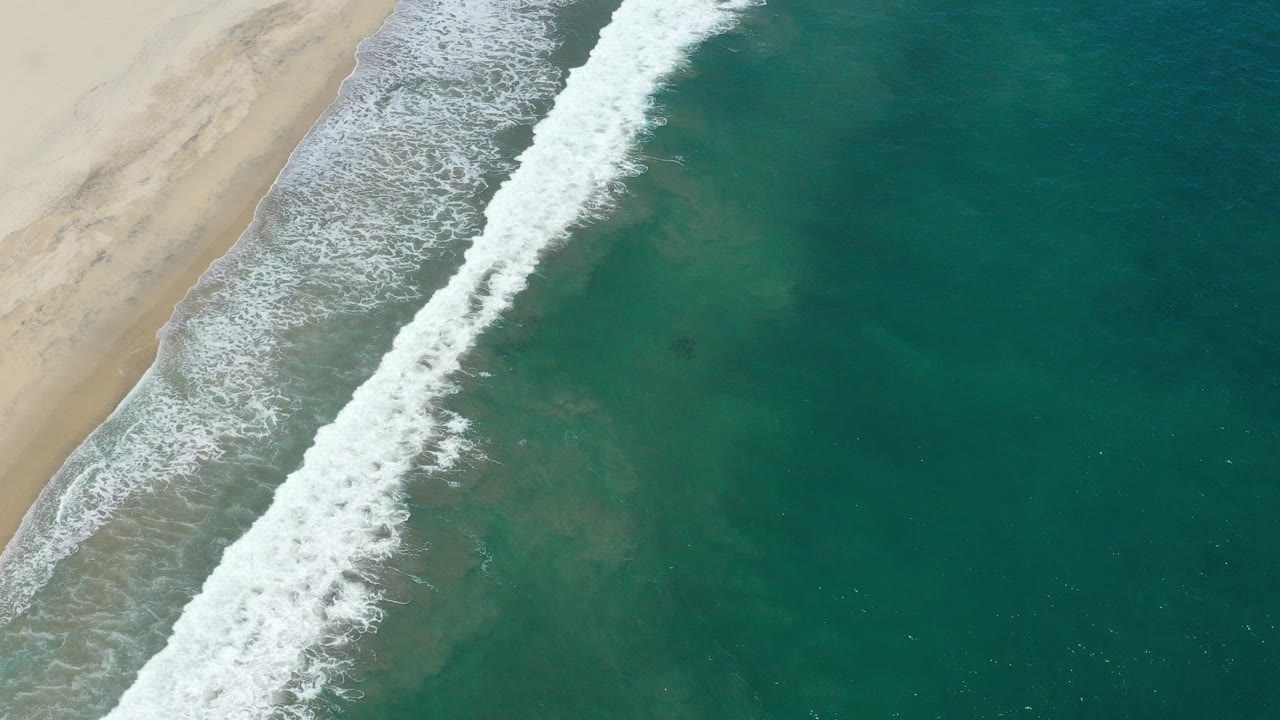Aerial view of white surf meeting pale sand and teal water in San Jose del Cabo, Mexico, with no visible people.