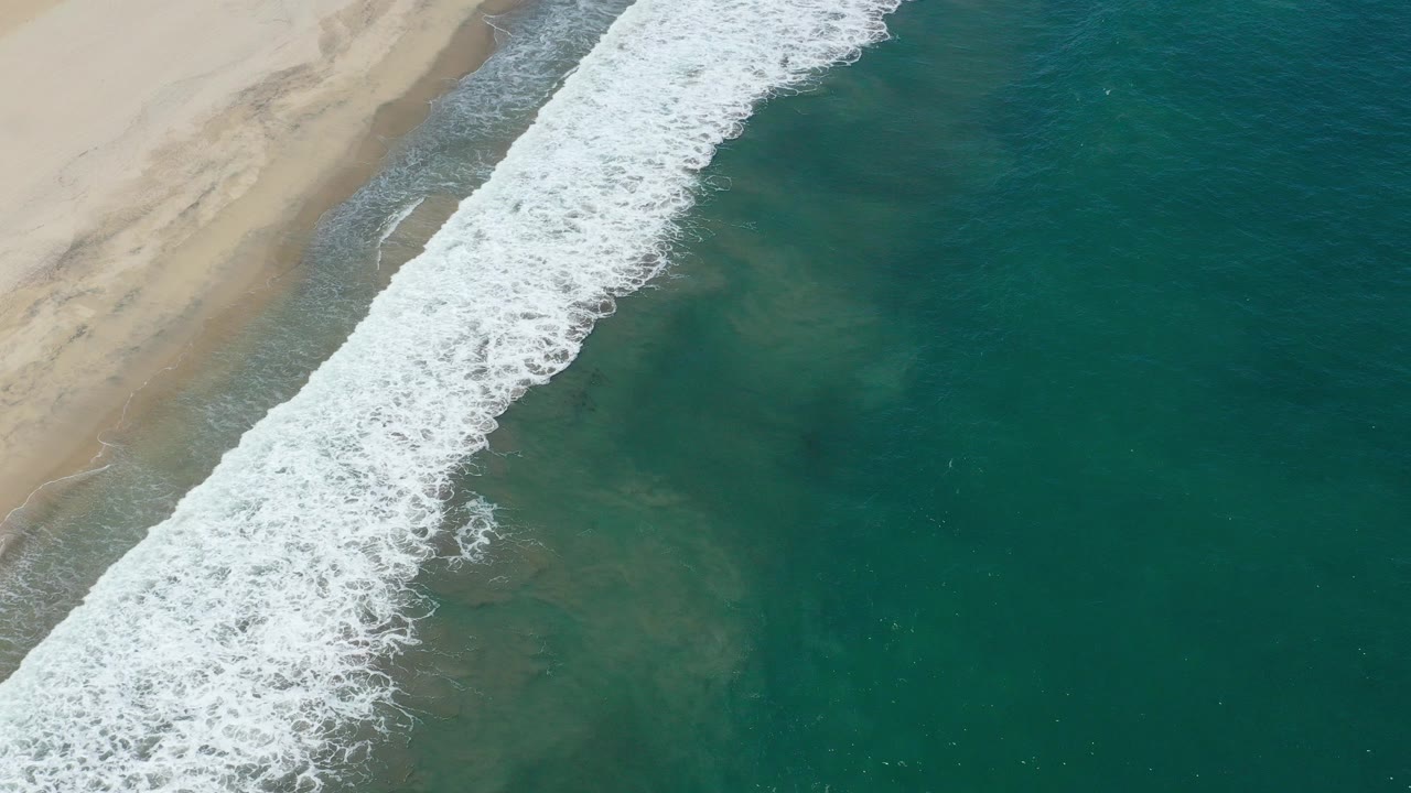 Aerial frame over San Jose del Cabo surf, with pale sand at the upper left, a diagonal band of white foam, green water to the right, and no visible people.