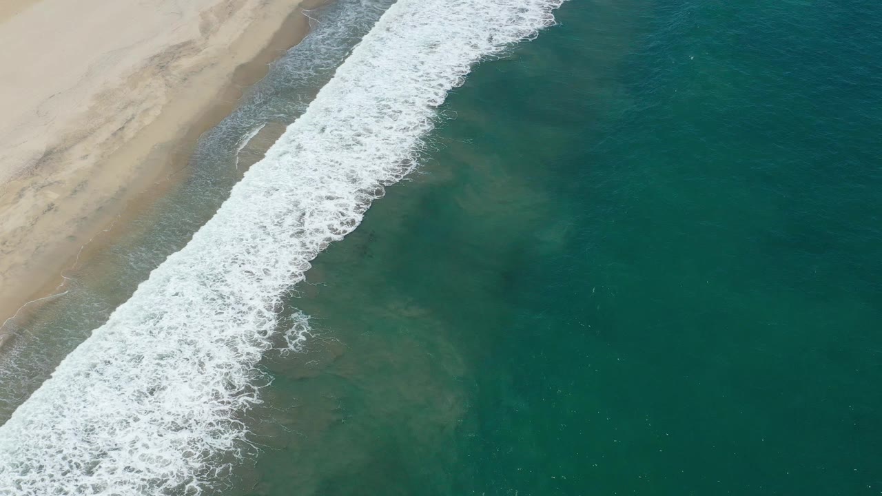 Aerial view in San Jose del Cabo with the foam line shifted slightly down the beach, translucent green water on the right, pale sand on the left, and no visible people.