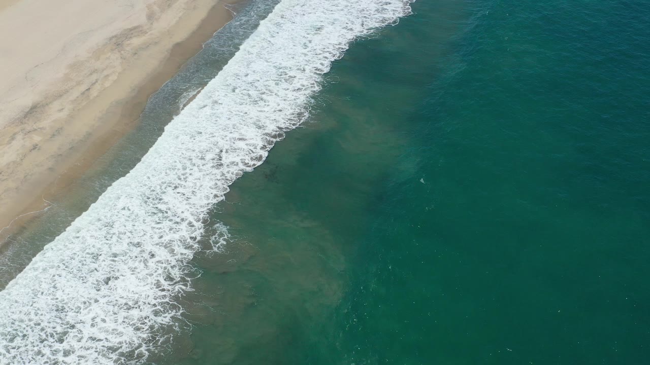 Breaking surf spreads along the edge of a sandy beach with open green water.