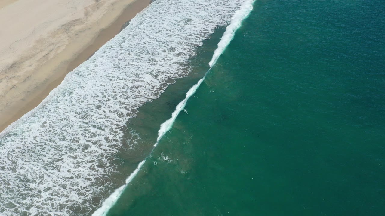 Aerial frame from San Jose del Cabo showing a diagonal line of white surf beside beige sand and teal water, with no visible people.
