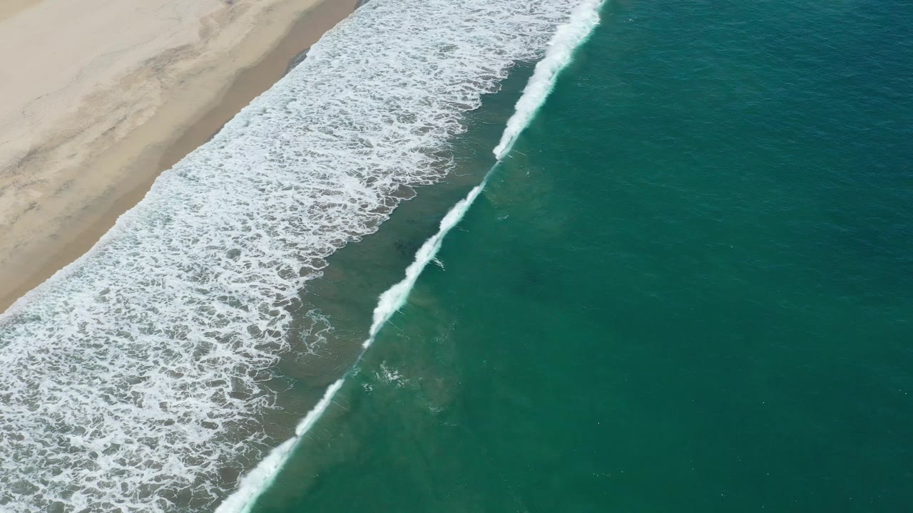 Aerial San Jose del Cabo shoreline frame with the foamy break shifted along the beach and open teal water filling the right side, with no visible people.