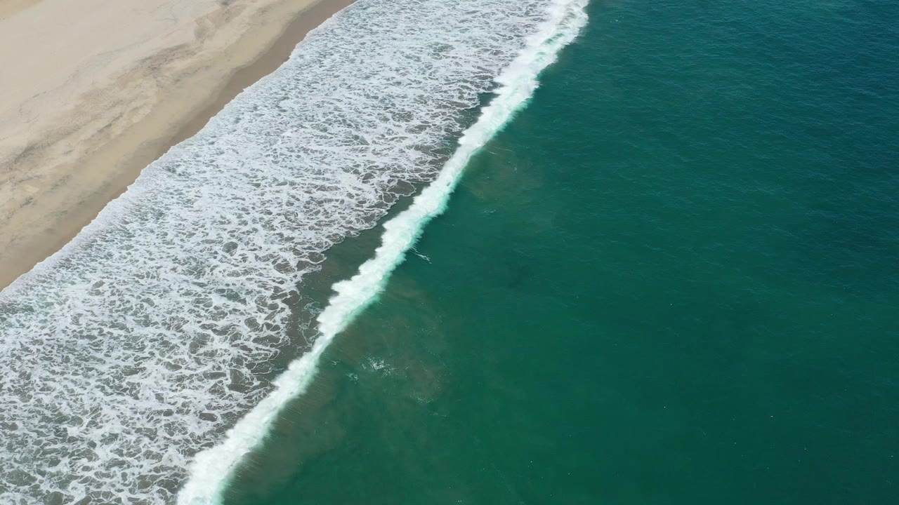 Aerial view in San Jose del Cabo with a brighter band of breaking white waves near the left side and broad teal ocean on the right, with no visible people.