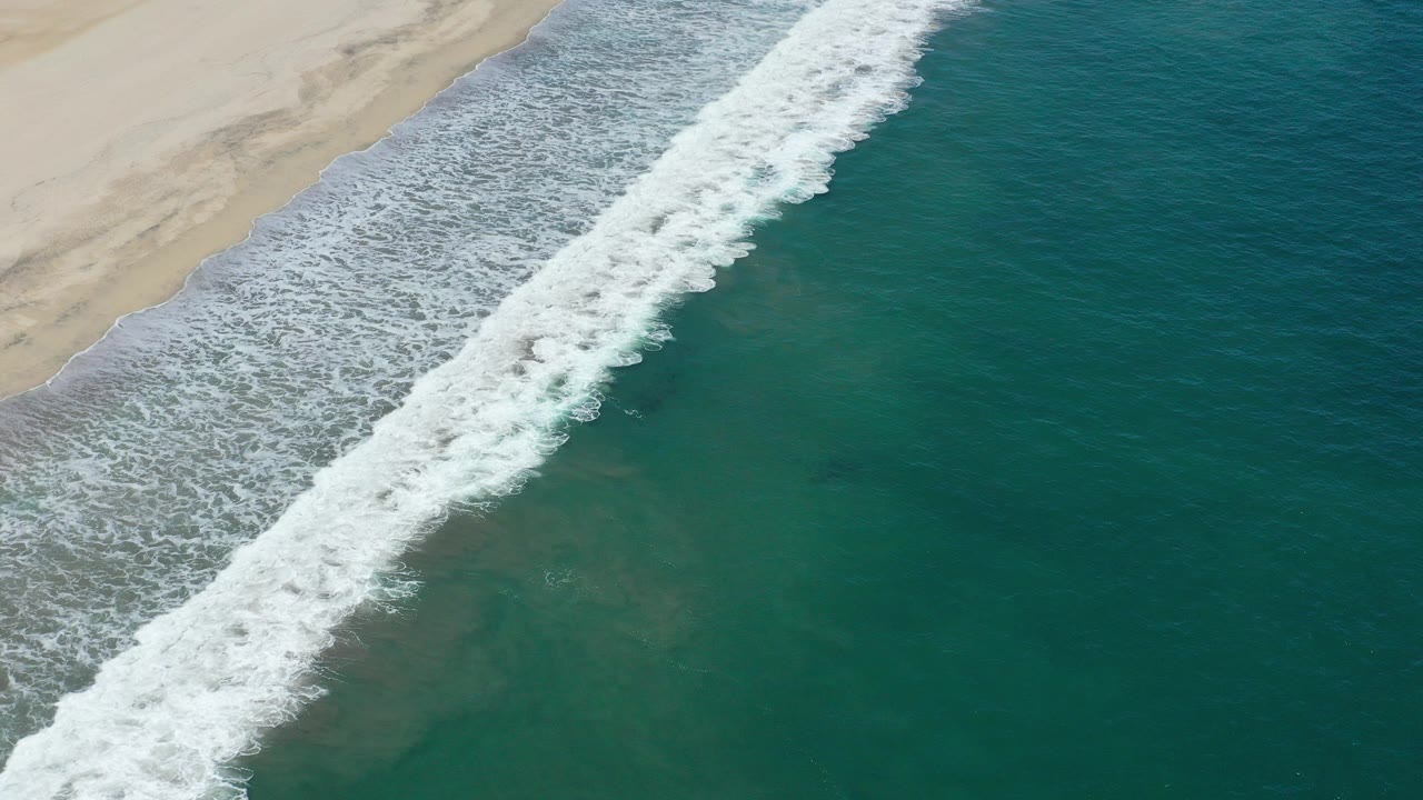 Overhead beach wave breaking beside teal water and pale sand.
