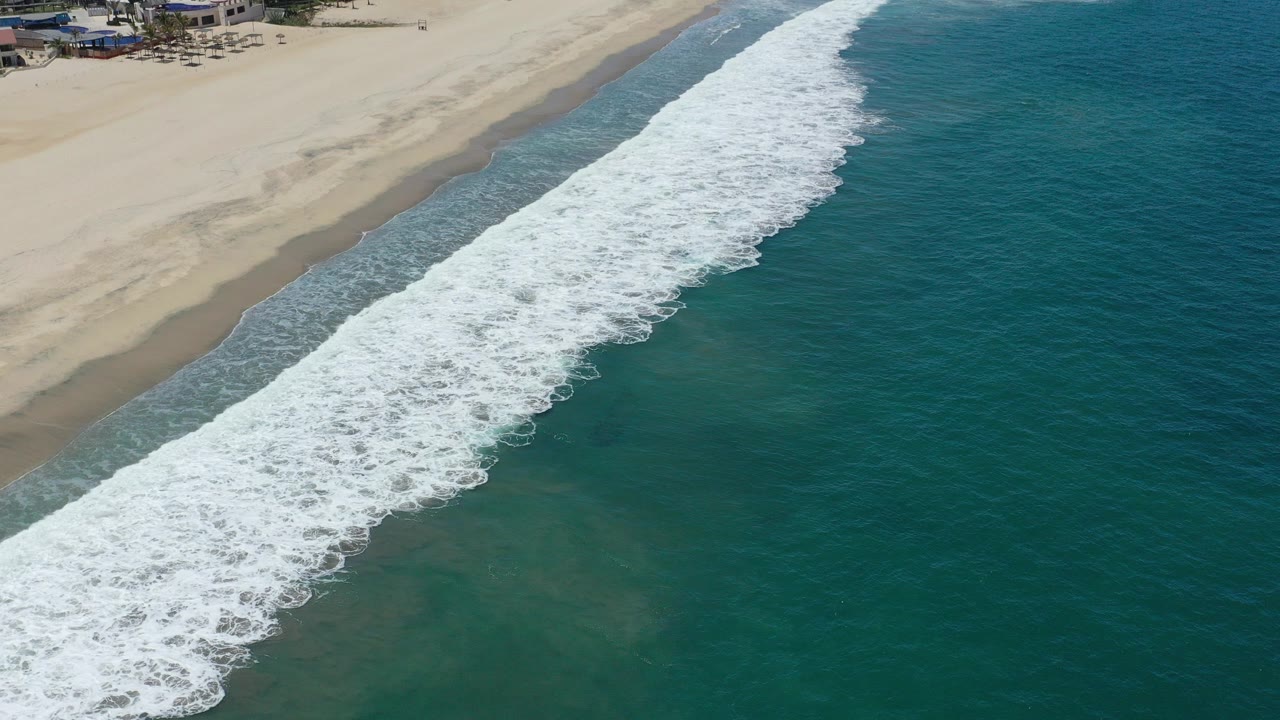 Aerial view of San Jose del Cabo surf forming a wide white diagonal band between teal ocean and tan sand, with distant beach buildings at the top edge and no clearly visible people.