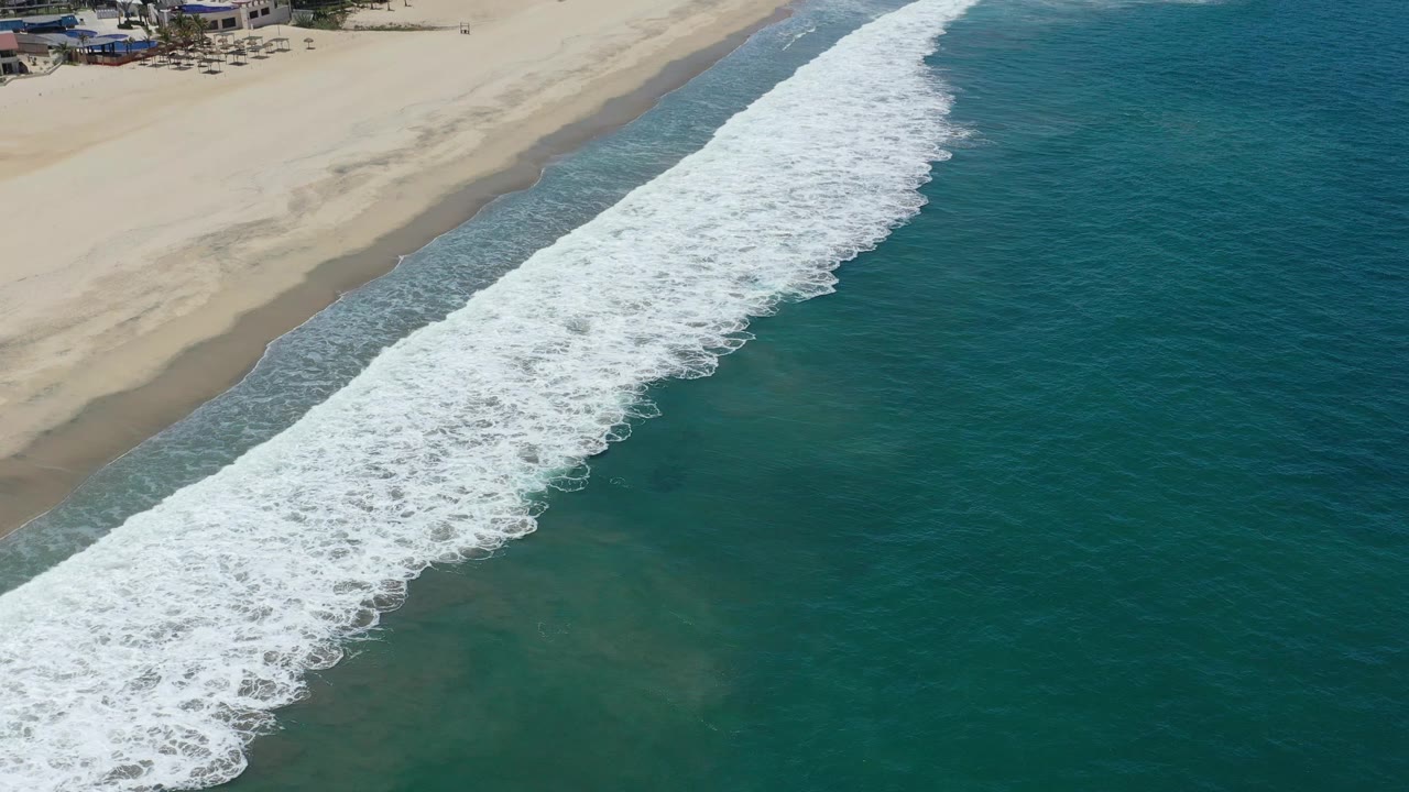 Aerial San Jose del Cabo shoreline view with the foamy breaker line shifted farther across the frame, teal water filling the right side and pale sand filling the left.