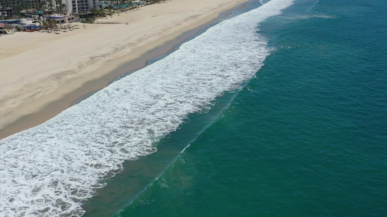 High aerial view over San Jose del Cabo, Mexico, with a broad white surf band curving between teal ocean and pale sand, with beachfront buildings at the upper edge and tiny distant people on shore.