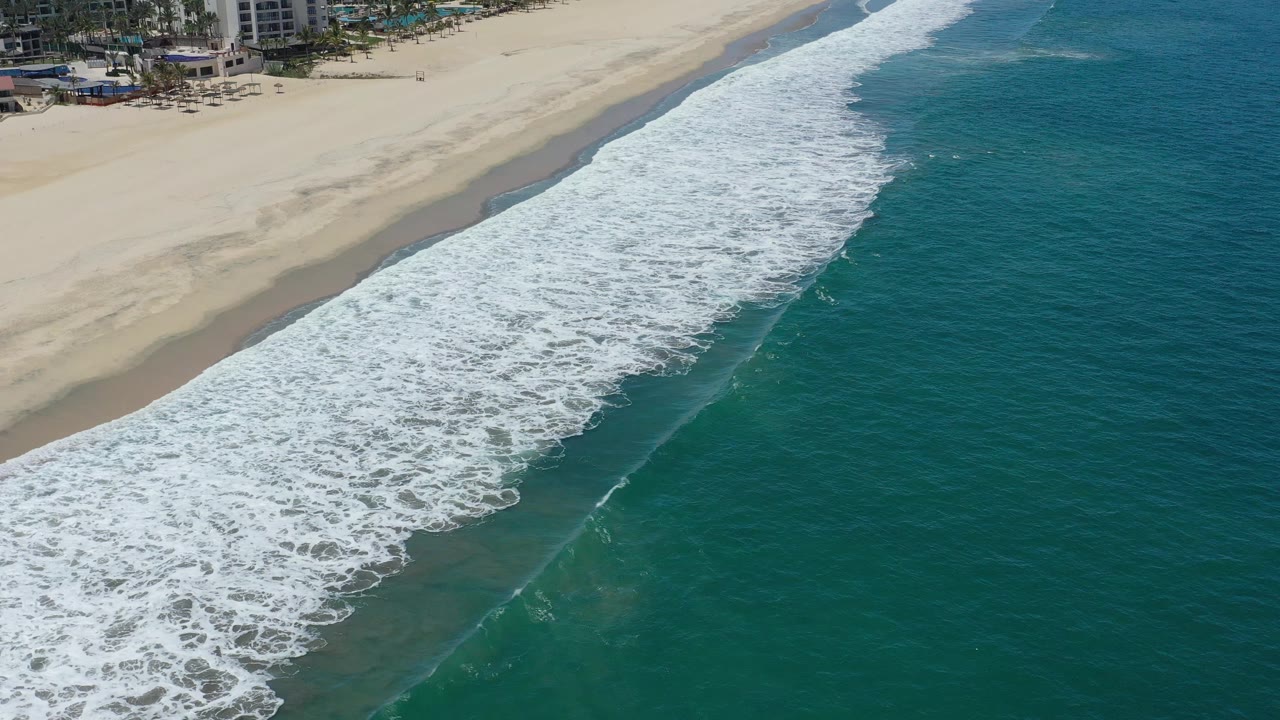 Aerial frame from San Jose del Cabo, Mexico, showing foamy white water stretched along the beach, darker teal ocean to the right, and resort-area structures near the top left.