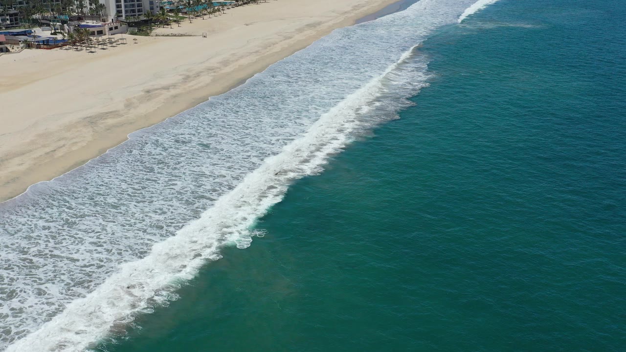Aerial frame from San Jose del Cabo showing green ocean on the right, a diagonal white wave breaking toward pale sand, partial beachfront buildings at the top edge, and no visible people.