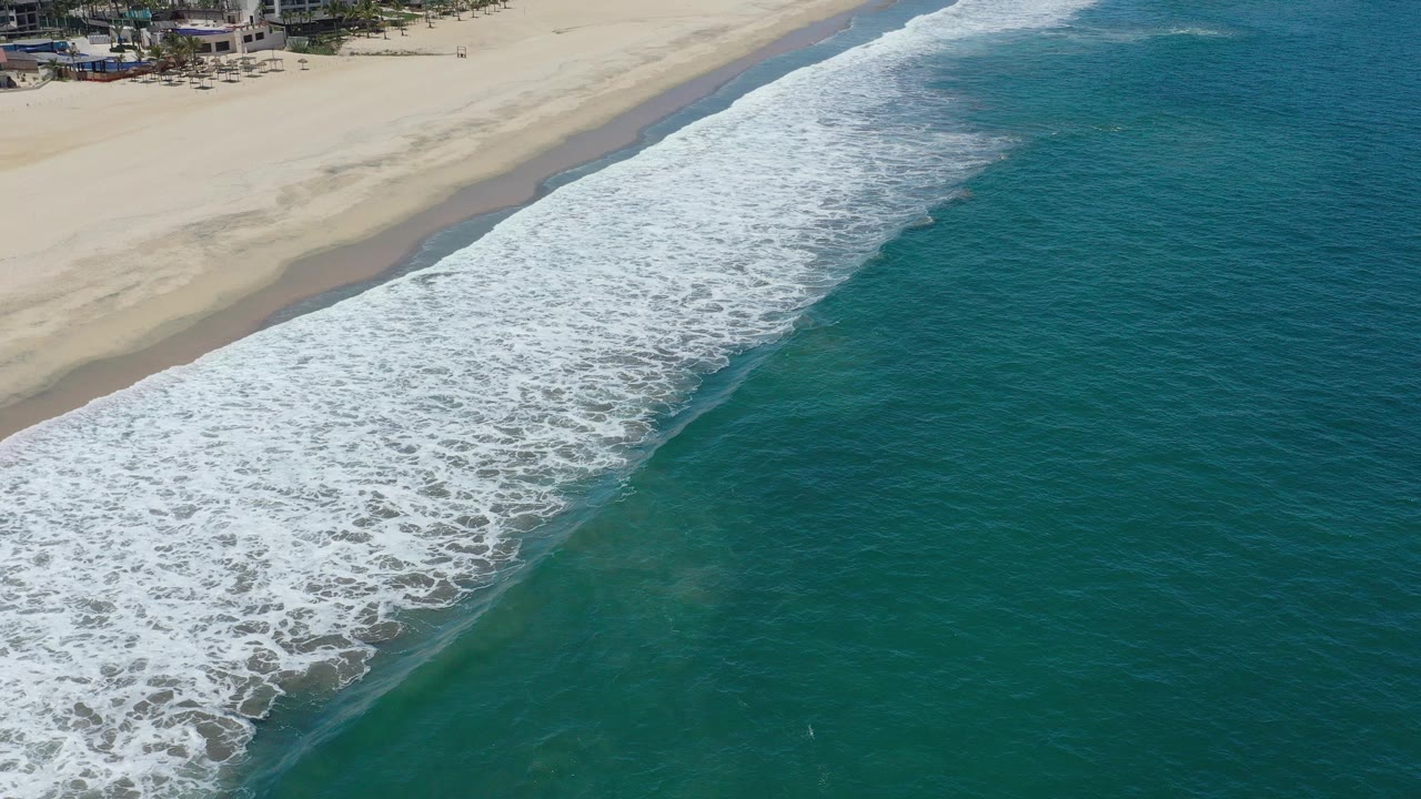 Aerial view in San Jose Del Cabo, Mexico, with white foam spreading across a diagonal shoreline, green-blue water on the right, pale sand at the top, and no visible people.