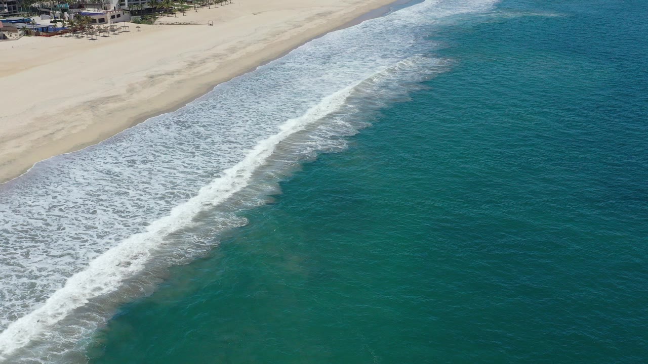 Aerial view of white surf meeting pale sand beside turquoise water.