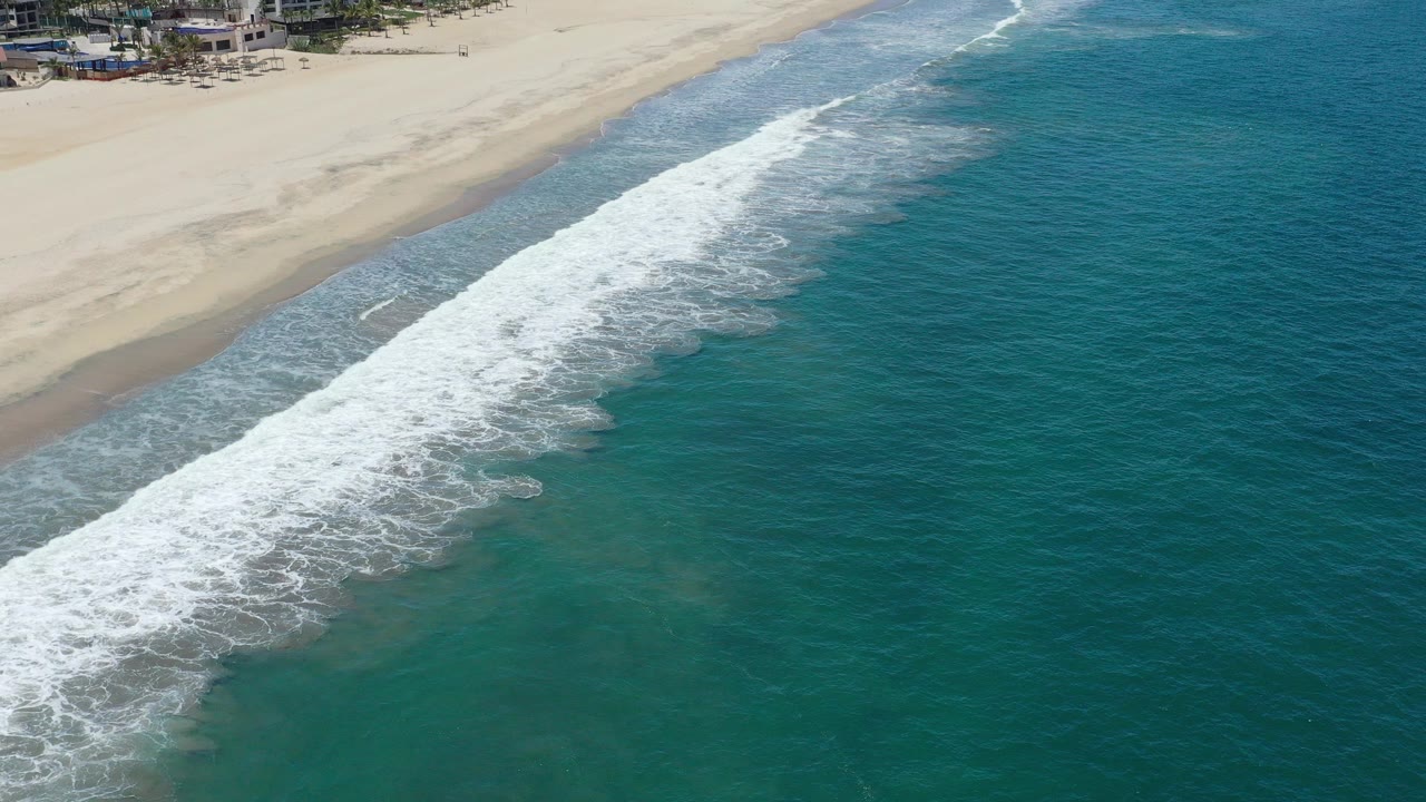 Aerial view of white surf running diagonally toward a sandy San Jose del Cabo beach, with teal water filling the frame and no visible people.