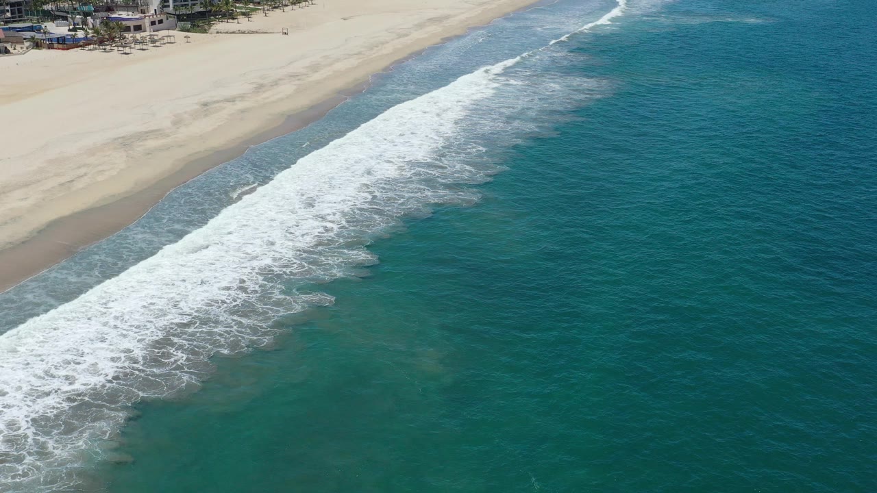 Breaking waves create a wide white band between turquoise ocean and sand on the San Jose del Cabo coast, with no visible people in the beach area shown.