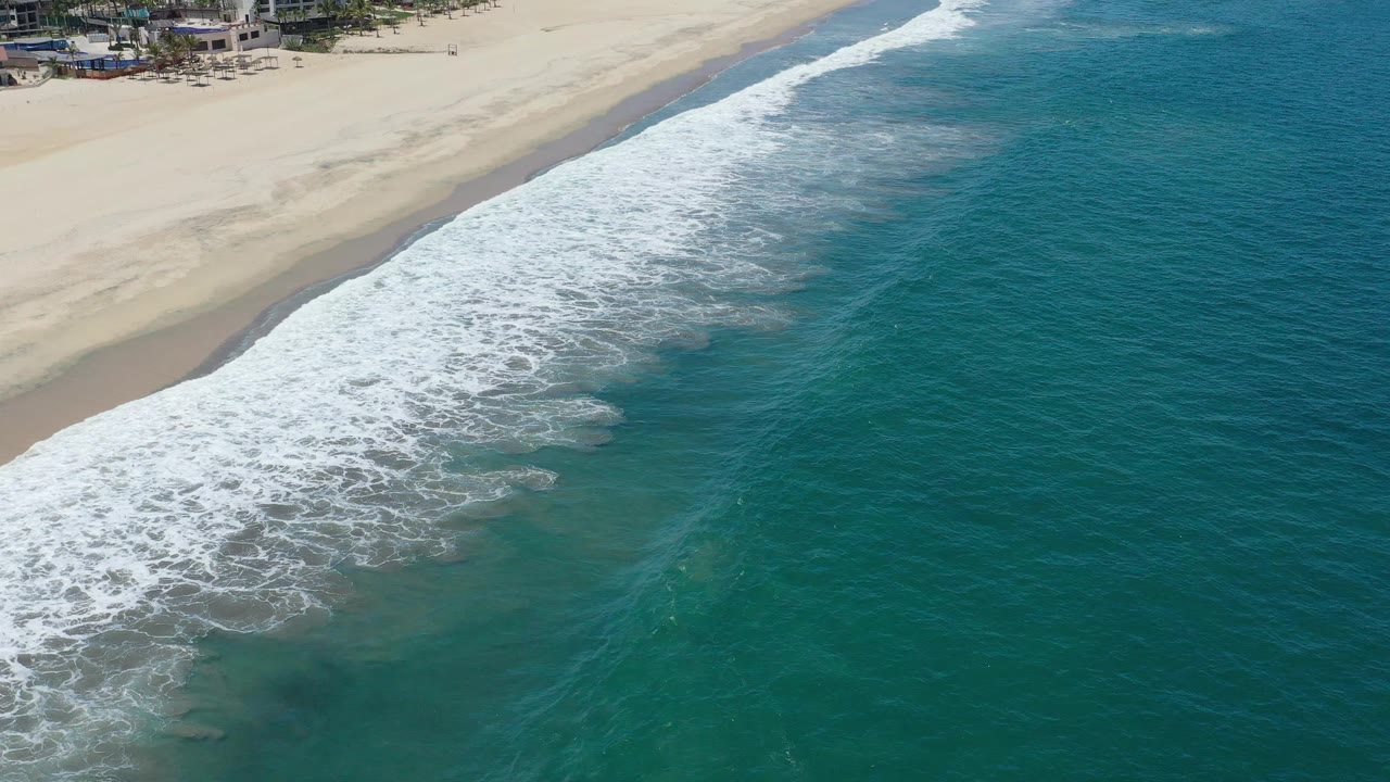 Aerial shoreline frame in San Jose Del Cabo, Mexico, showing foamy waves spreading farther along the sand with open blue-green water beside them.