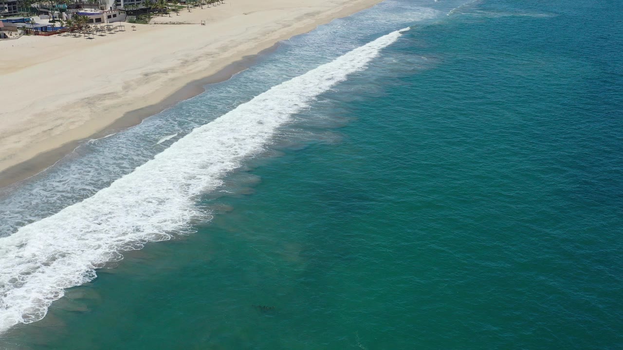 Opening aerial frame over San Jose del Cabo, Mexico, with white surf running diagonally across green sea toward pale sand and no visible people.