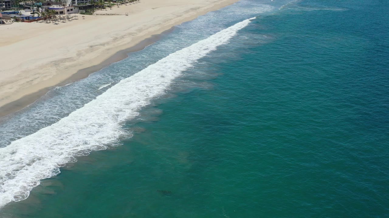 Aerial frame over San Jose del Cabo, Mexico, where a broad white wave band spreads along the beach edge with green water filling the foreground and no visible people.