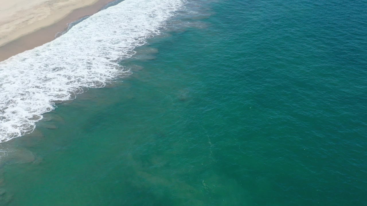 Aerial view over San Jose del Cabo shoreline with white surf curling along the sandy beach at the upper left and teal water below, no visible people.