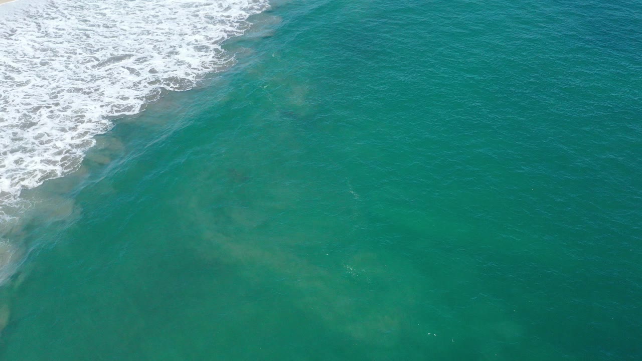 Aerial view of turquoise water in San Jose del Cabo, Mexico, with white surf entering from the upper left and pale current clouds under the surface, no visible people.