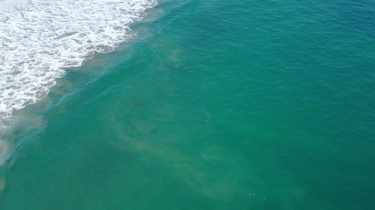 Aerial frame over San Jose del Cabo water where the white breaker line sits near the upper left and a broad pale current plume spreads through green-blue ocean, no visible people.