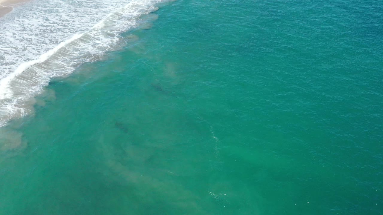 Aerial San Jose del Cabo water view with the foamy wave band sliding along the upper left and cloudy sand plumes beneath the teal surface, no visible people.