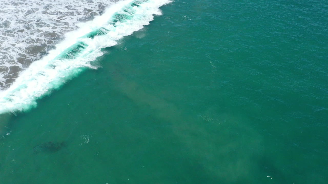 Aerial frame from San Jose Del Cabo showing turquoise water, white surf entering diagonally from the upper left, faint fish-like shapes below the surface, and no visible people.